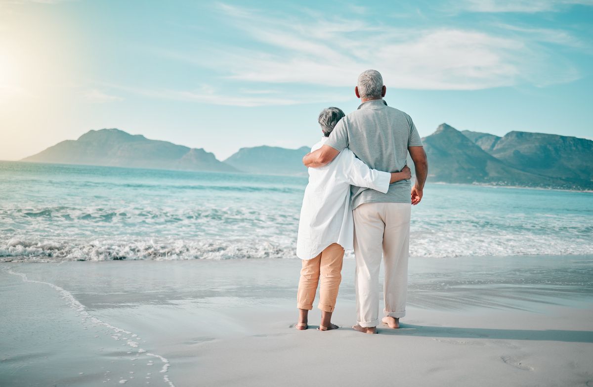 Two people standing arm in arm on a beach, facing turquoise water and distant mountains