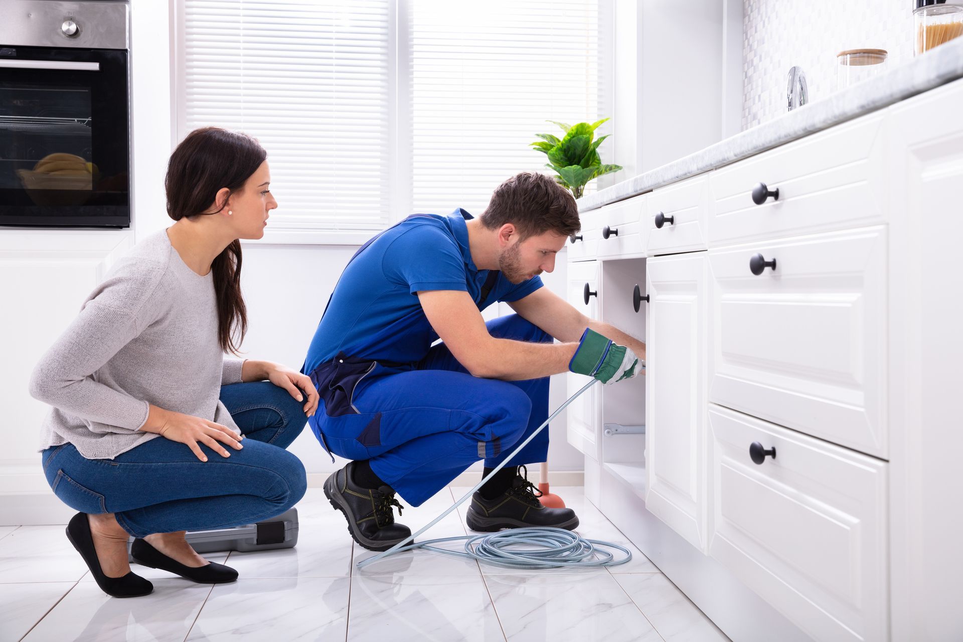 Plumber fixing kitchen sink pipe while homeowner observes in modern white kitchen.