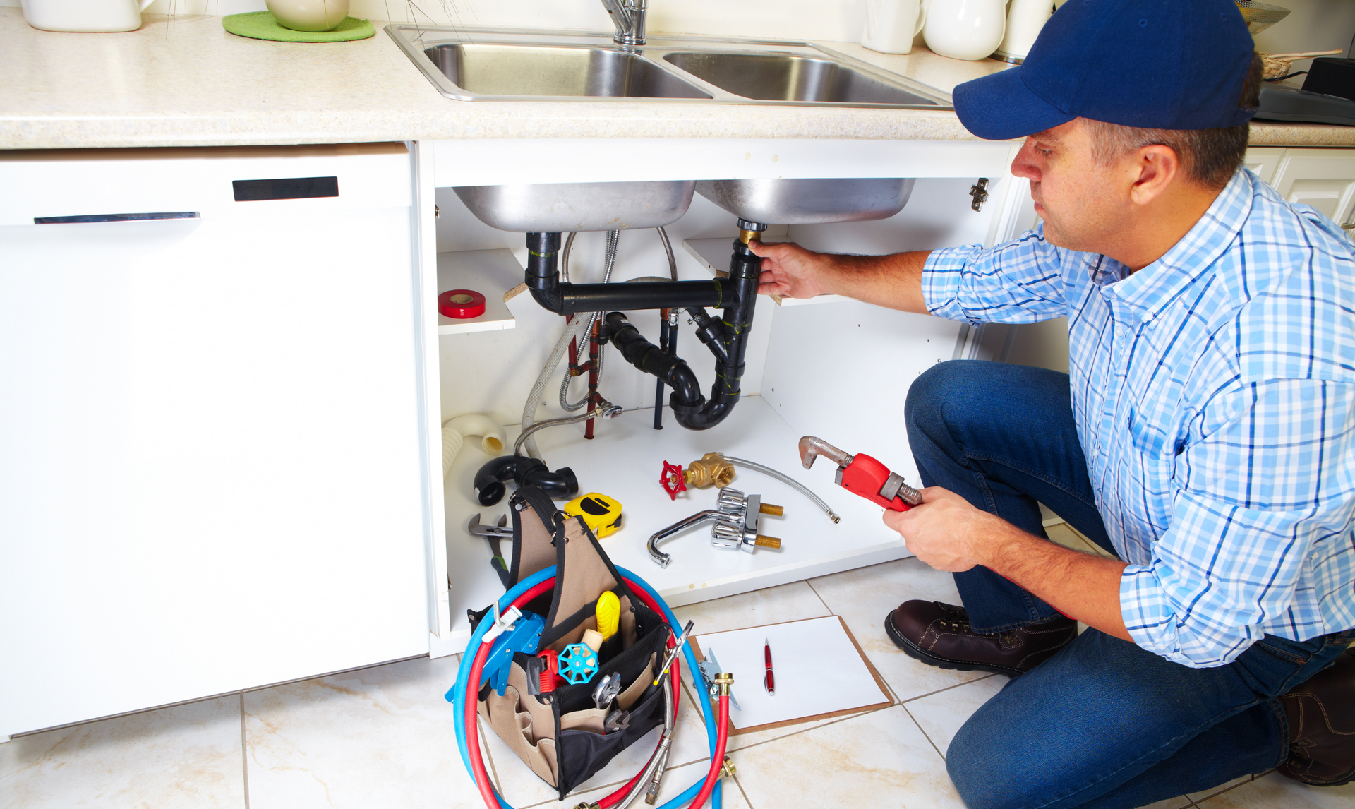 Plumber in a blue cap and plaid shirt repairs sink pipes, kneeling with tools and a toolbox under the sink. Plumber in a blue cap and plaid shirt repairs sink pipes, kneeling with tools and a toolbox under the sink.