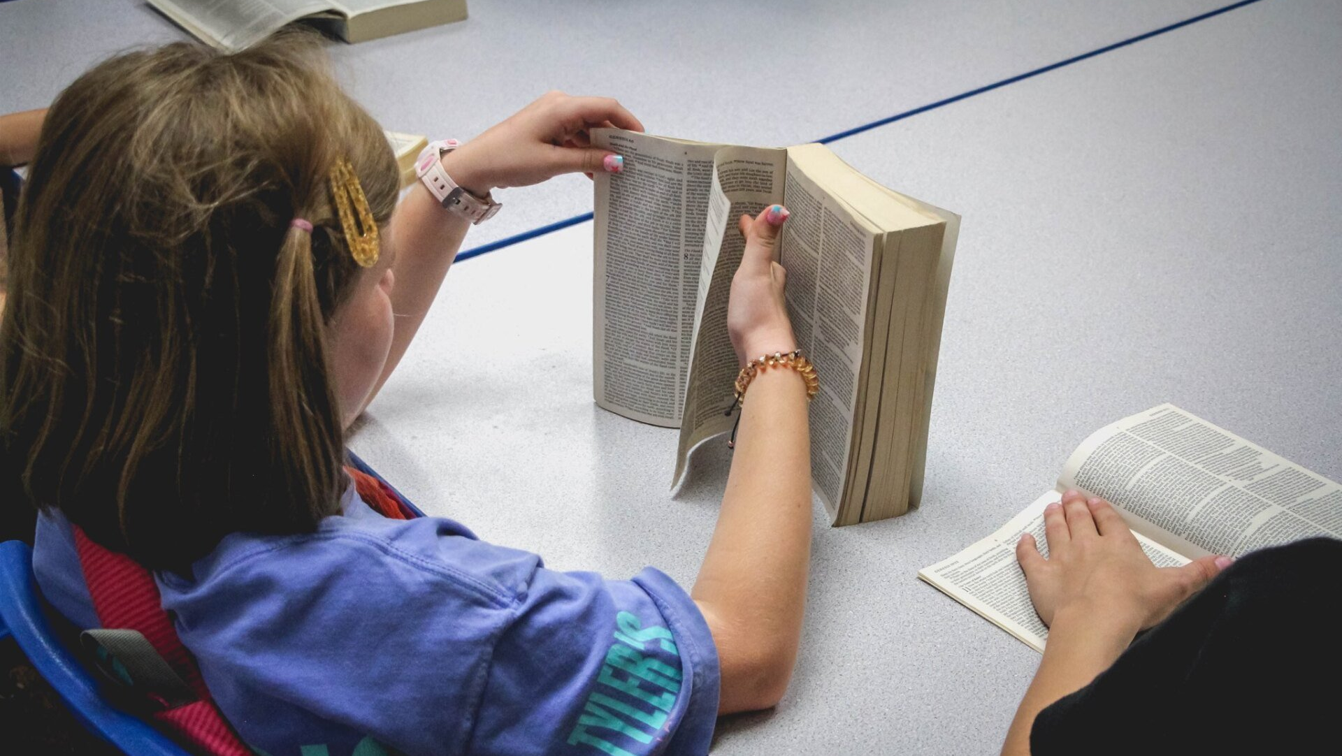 A child reading from the Bible, seated at a table with other books nearby.
