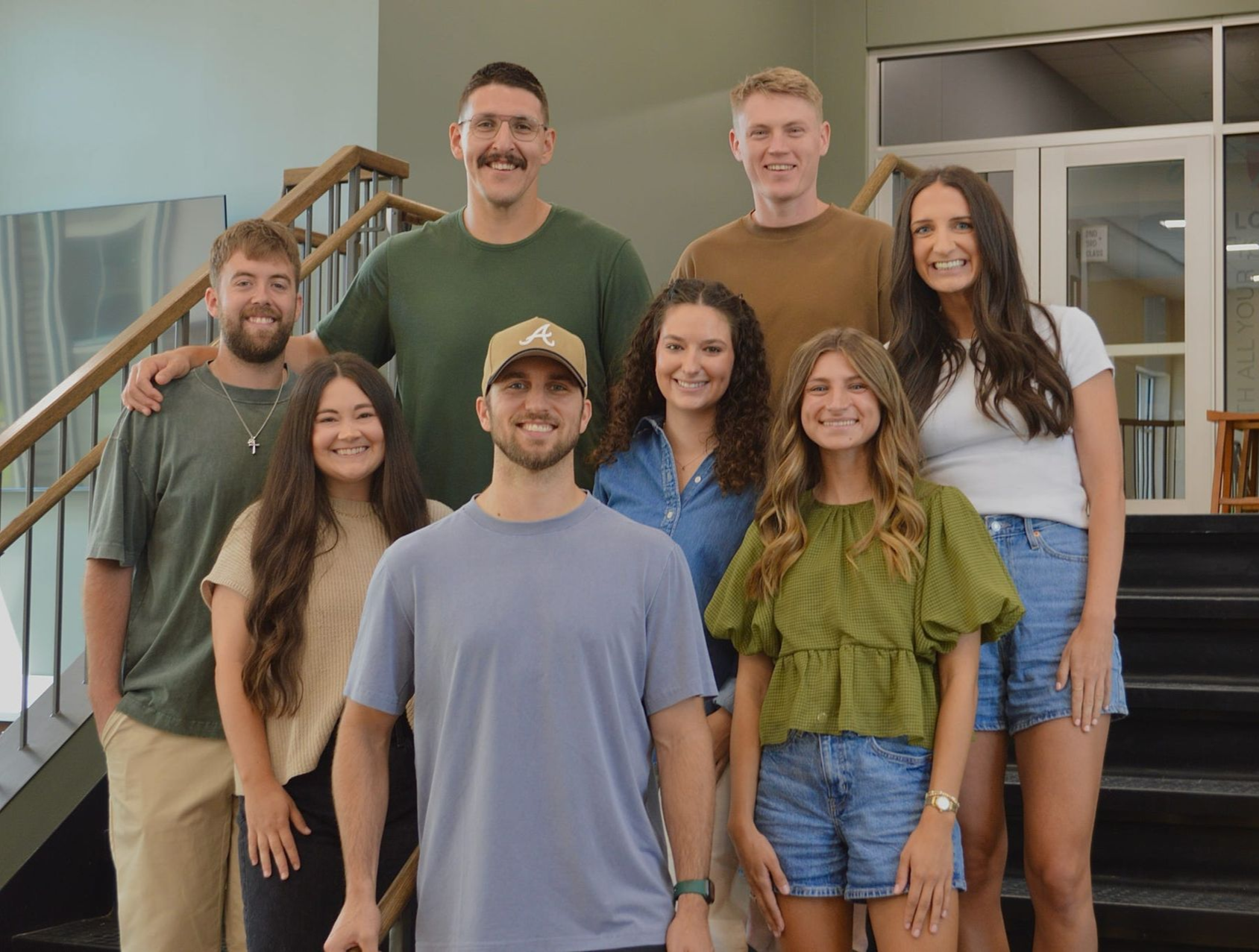 Seven smiling coworkers pose on office stairs and landing, wearing casual clothes.