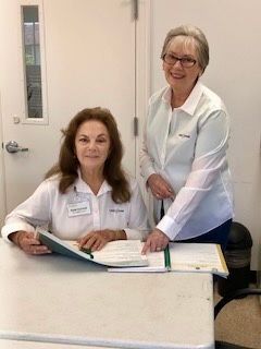 Two women at a table with papers, one seated, the other standing and pointing. Both wear white shirts and smile.