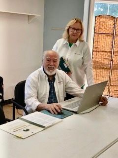 Man and woman at a table; man using a laptop, woman standing behind him holding a binder, both smiling.