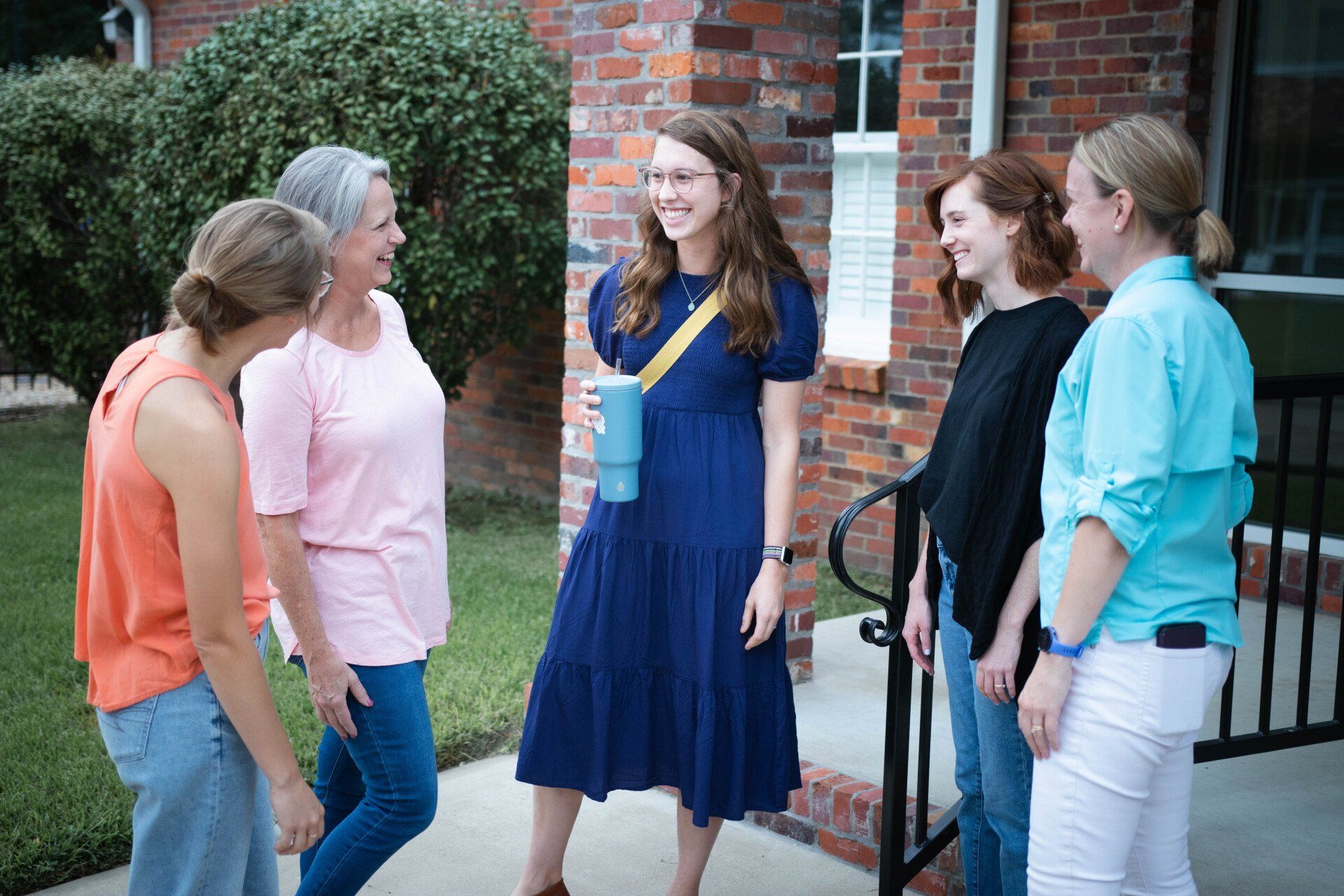 Group of people chatting on a porch. Woman in blue dress smiles, holding a drink. Others look on, laughing.