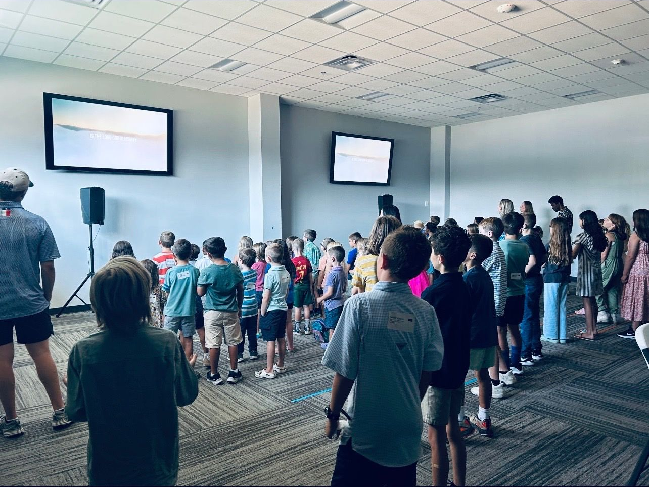Worship band performing on stage in the GBC sanctuary, audience of children watching from below, projection on the wall.