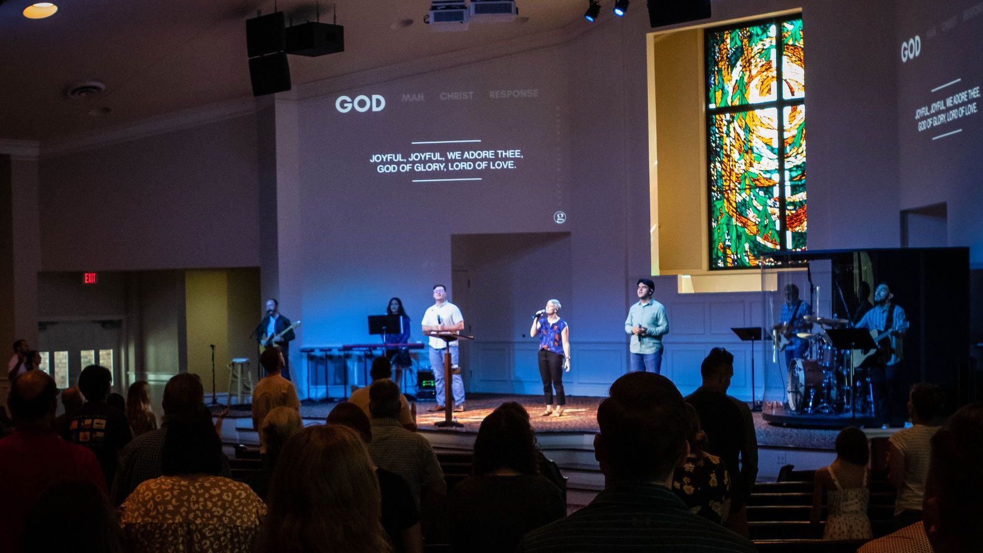 Worship at GBC, stage with band and singers, projected text, audience facing the stage.