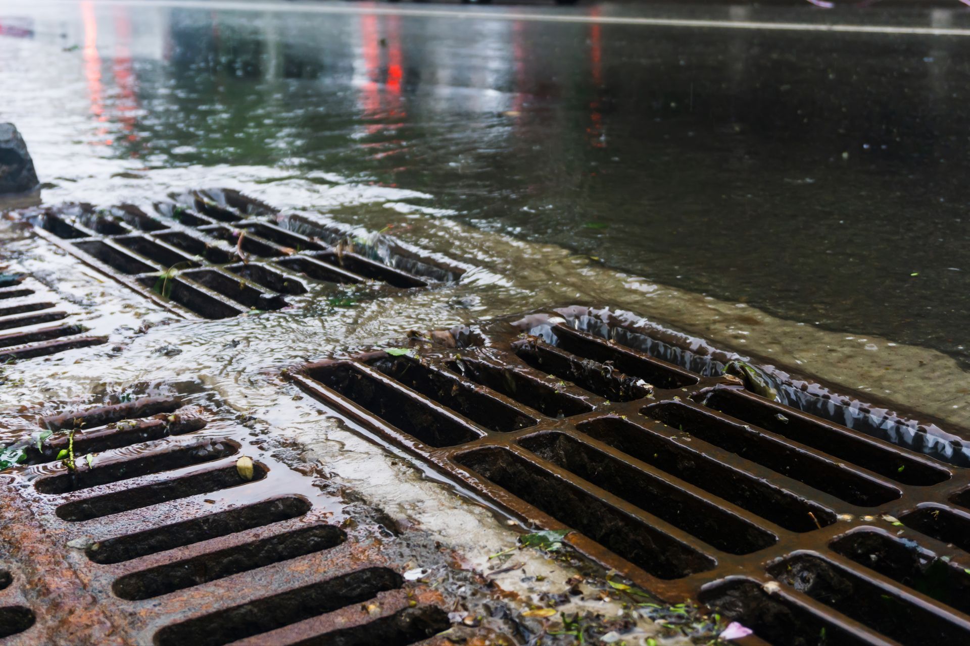 Photograph showing water flowing into rusty metal storm drains on a wet street surface.