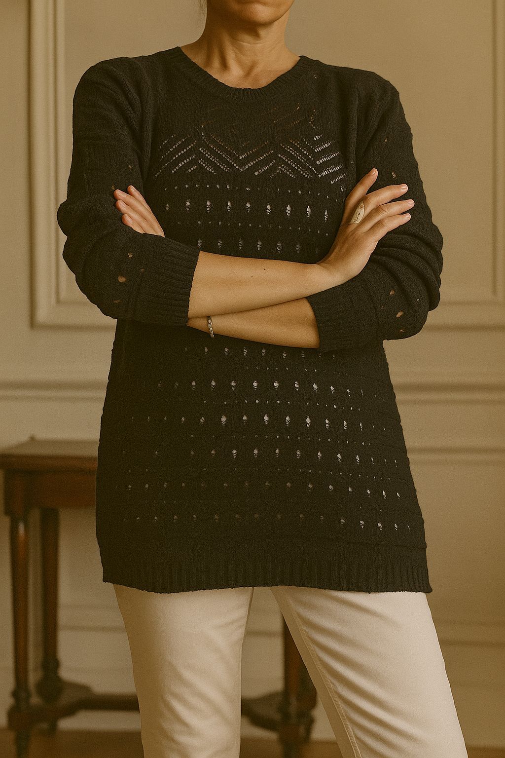 Mujer que viste un suéter oscuro con detalles calados, con los brazos cruzados, de pie en una habitación.