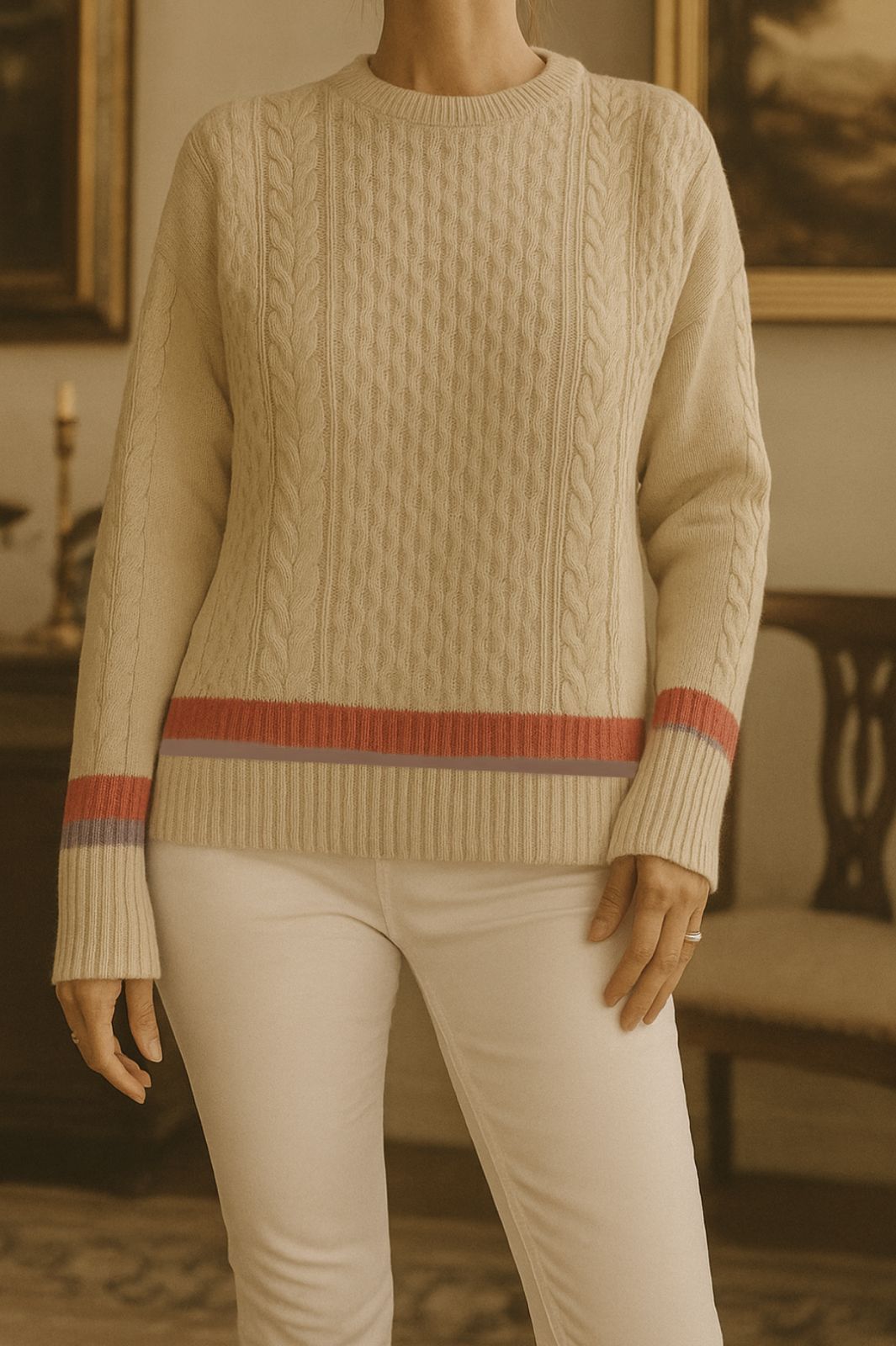 Mujer con suéter de punto blanco roto con rayas rojas y violetas, pantalones blancos, de pie en el interior.