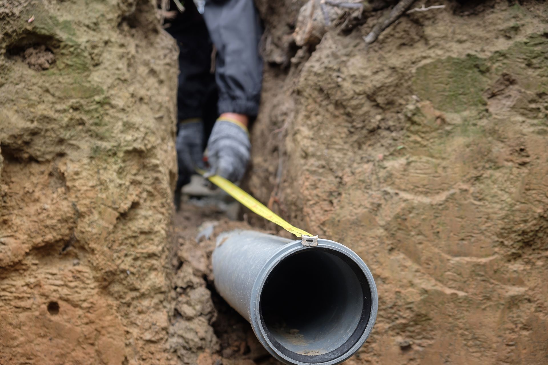 A worker measuring a large gray pipe in a dirt trench with a tape measure.