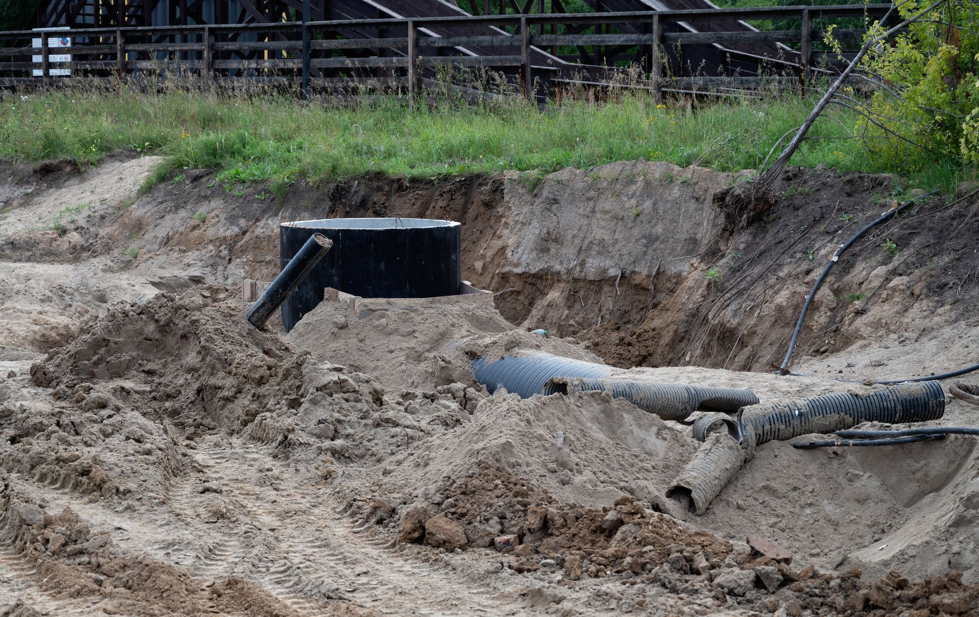 Mud-filled excavation site with exposed pipes and a black drainage opening along a fence line.
