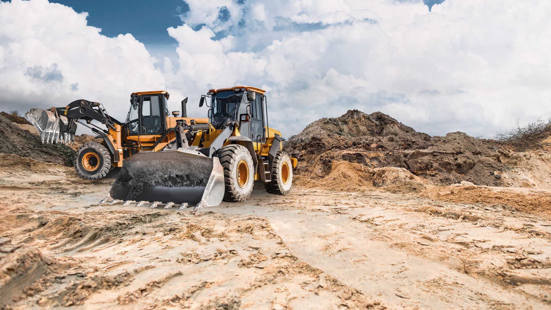 Two yellow front loaders on a sandy construction site.