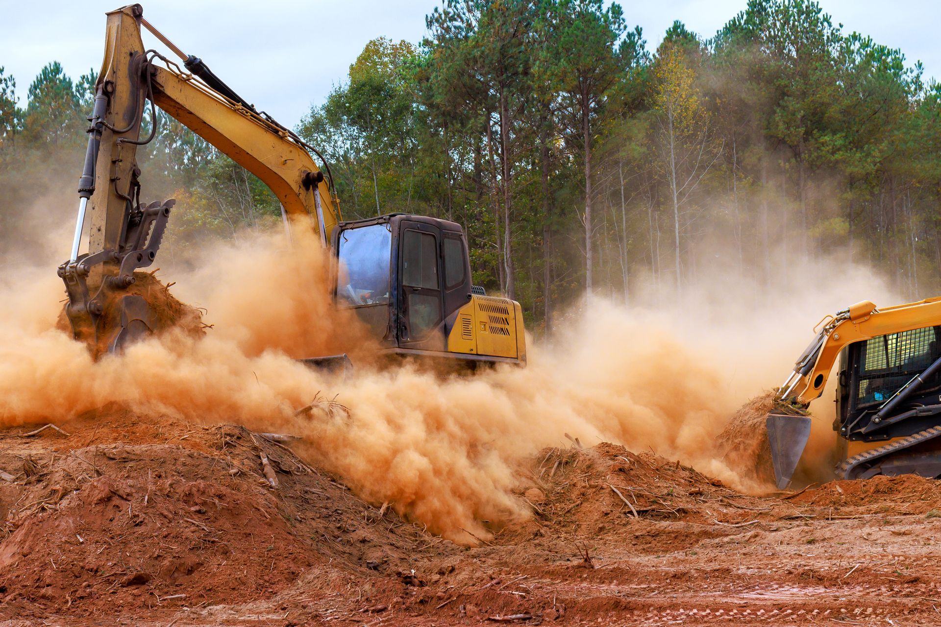 Two excavators kicking up a huge dust cloud at a dirt worksite in a wooded area.