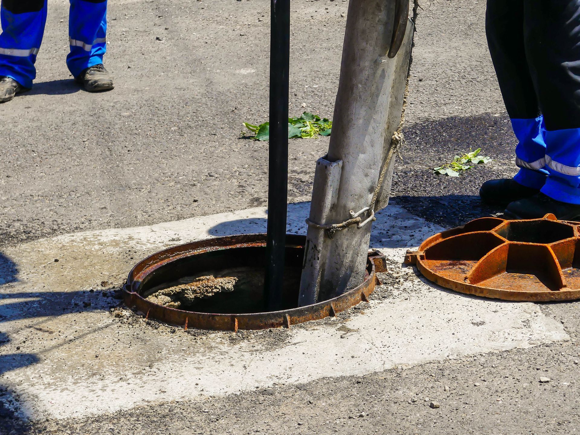 Workers in blue uniforms beside an open manhole with a metal pole on a street curb.