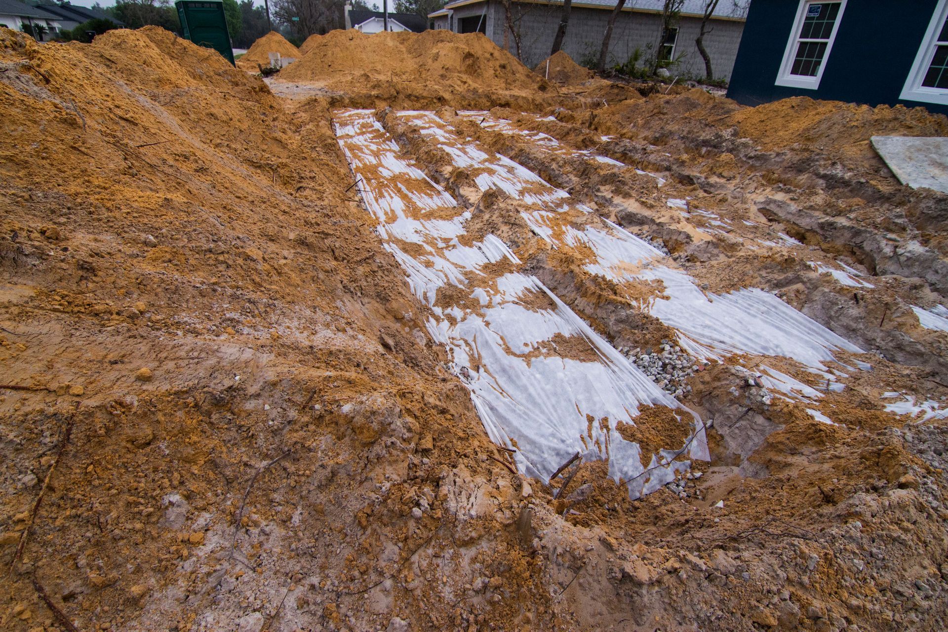 Muddy construction site with dirt piles and a white tarp-covered area beside a house.