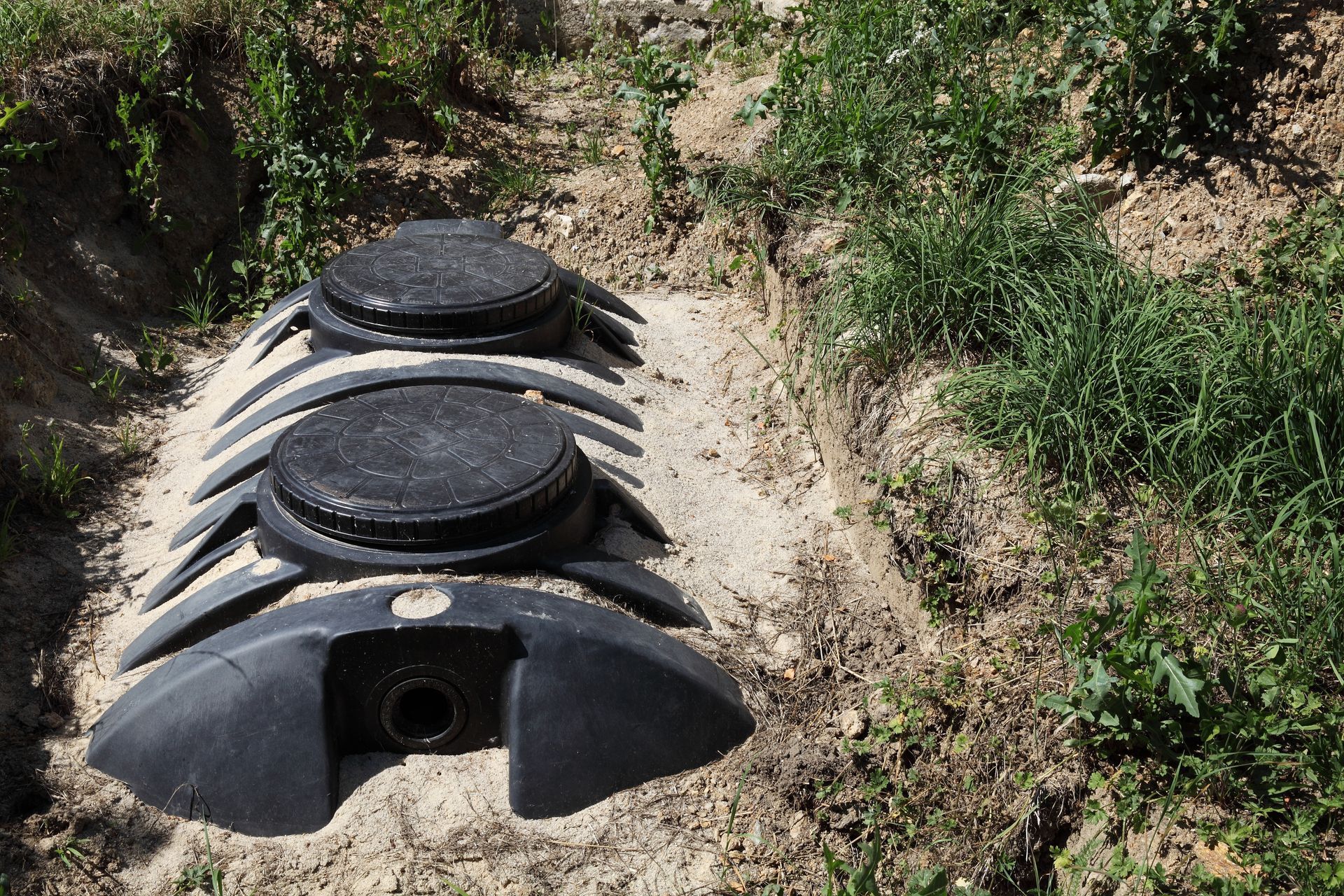 Worker using a corrugated hose to clean a grass-covered drain opening outdoors.