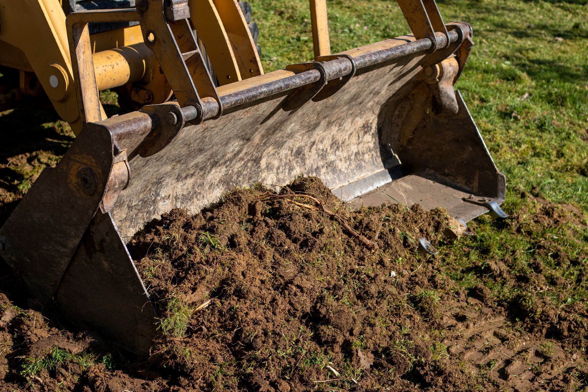 Excavator bucket scooping soil from a dirt pile on a grassy site.