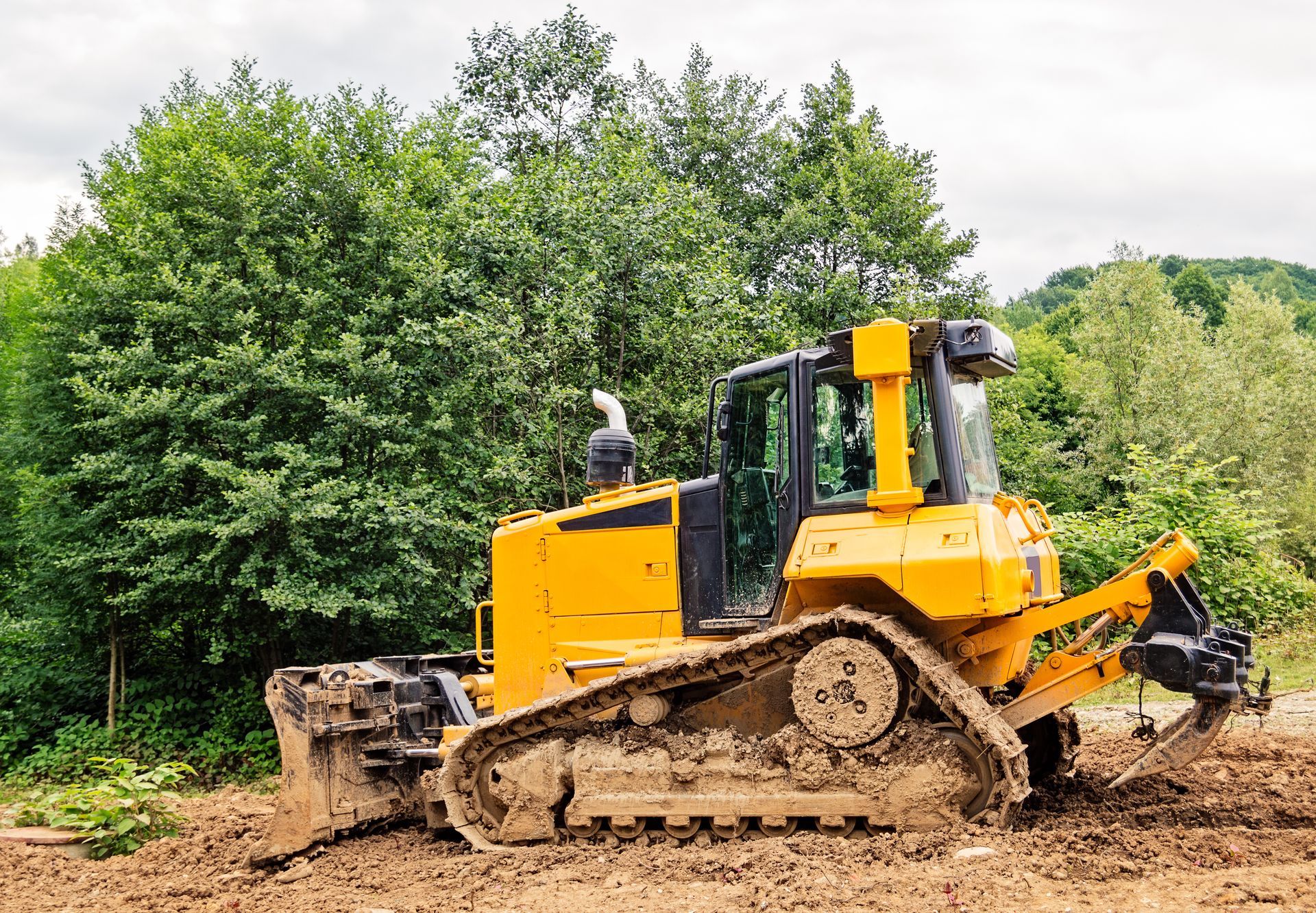 Yellow bulldozer on muddy ground near green trees and a cloudy sky.