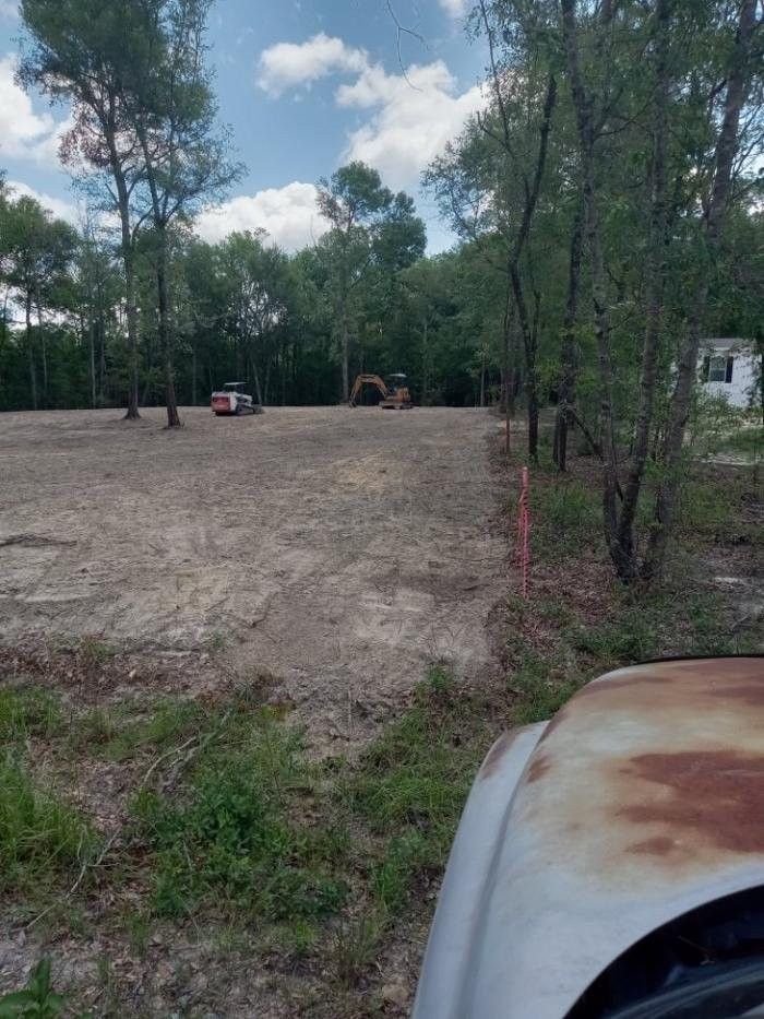 Dirt clearing in a wooded area with a red vehicle in the distance and a rusty white truck hood in the foreground