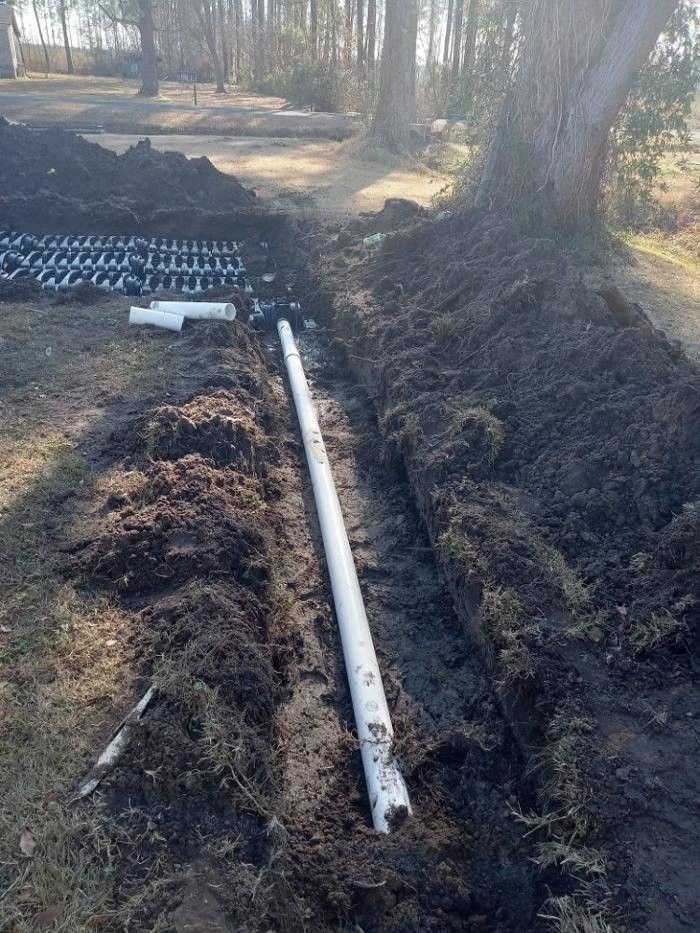 Utility trench with a white PVC pipe laid in dark soil beside a wooded path.