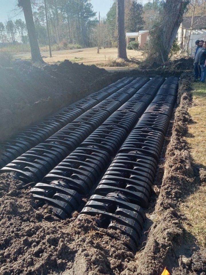 Black corrugated drainage pipes laid in a trench beside a dirt road in a rural area.