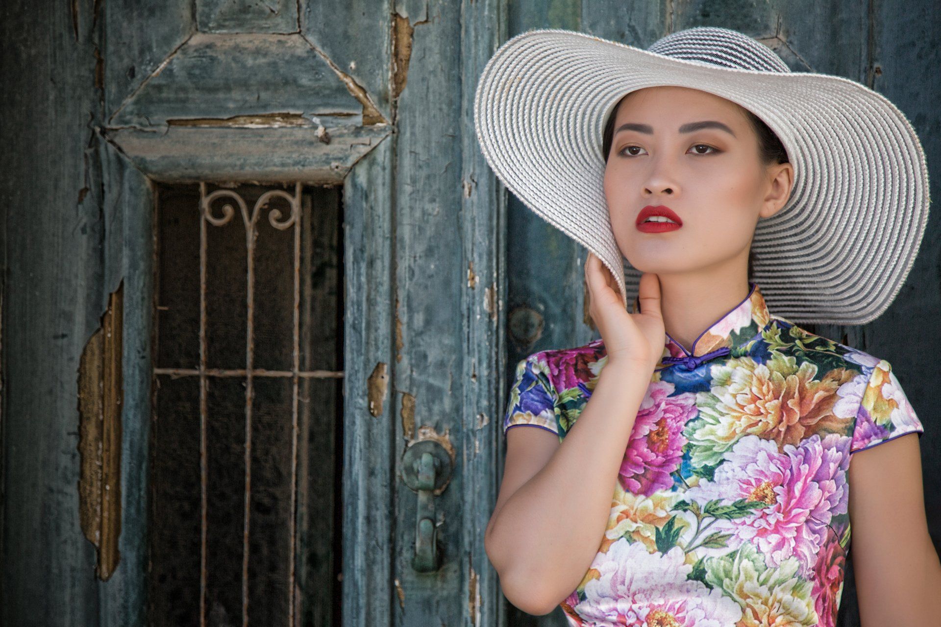 A woman wearing a hat and a cheongsam is standing in front of a blue door.