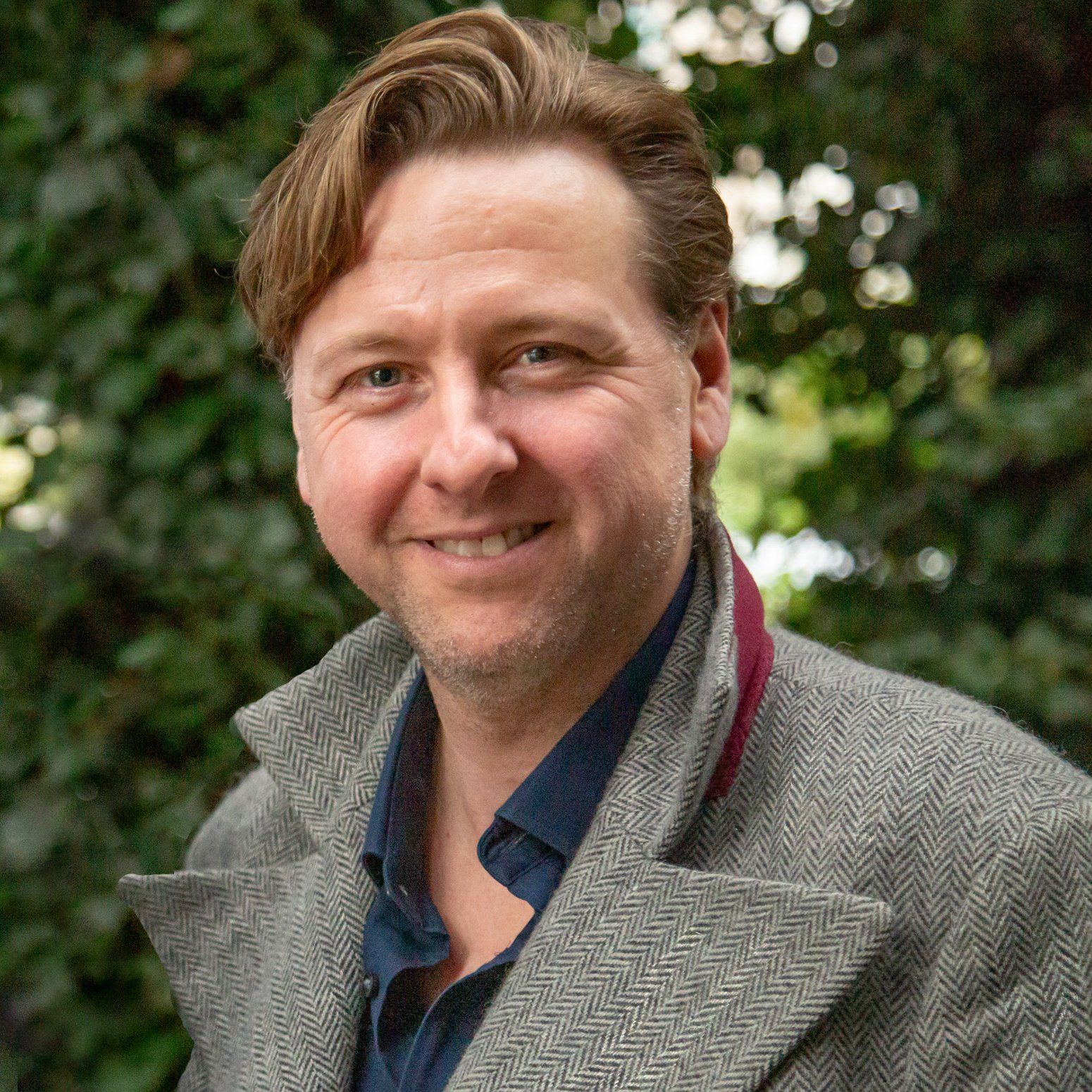 A man wearing a grey coat and a blue shirt is smiling for the camera.