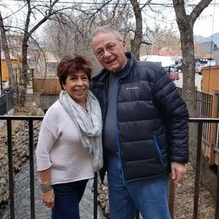 Two people standing together outdoors in front of a fence, wearing jackets and smiling.