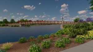 A landscaped riverfront with flowers and shrubs, a wide river, and a row of houses under a blue sky with clouds