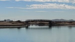 Wide calm river with a small bridge and distant mountains under a clear blue sky