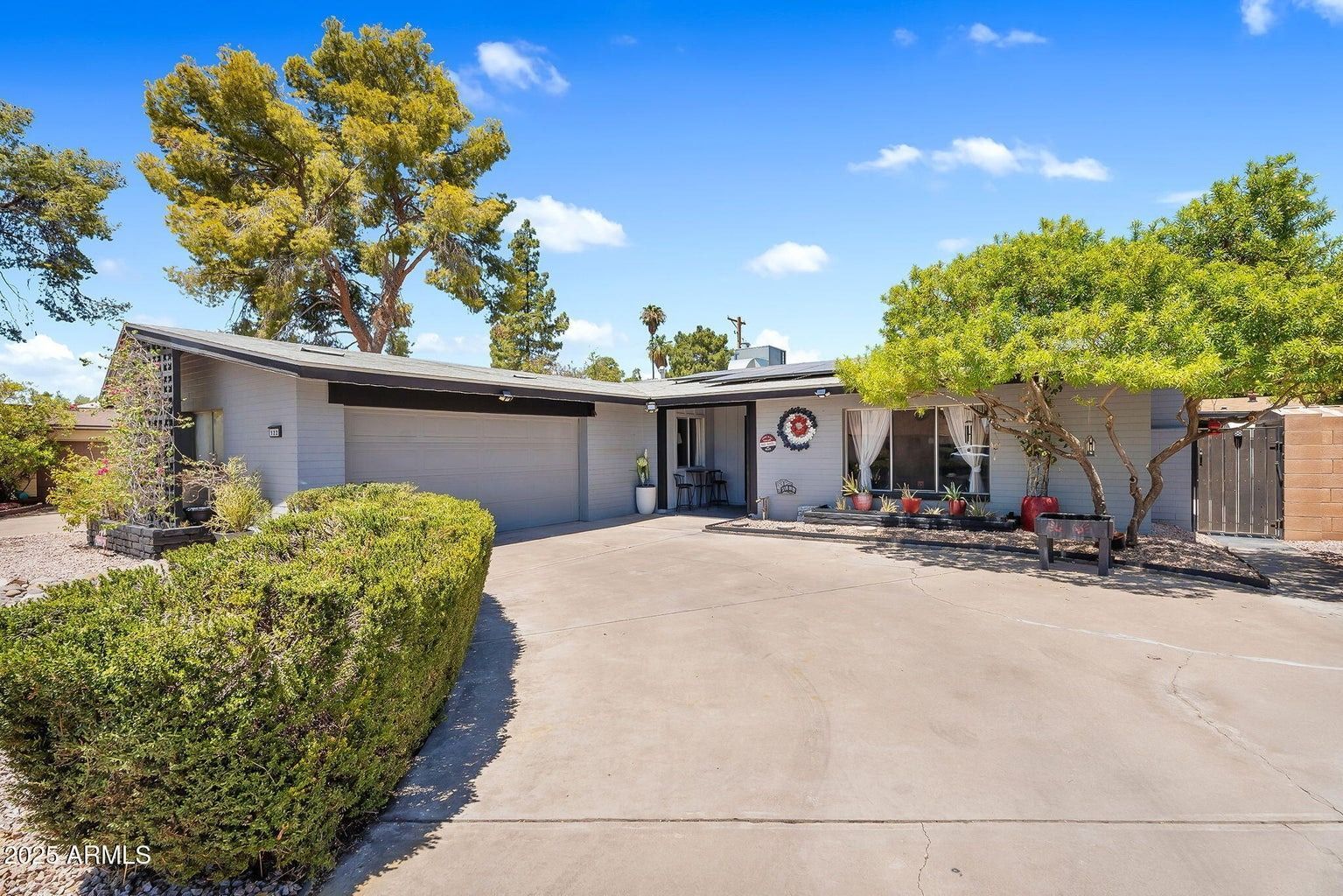 Single-story stucco house with a wide driveway, garage, desert landscaping, and trees under a blue sky