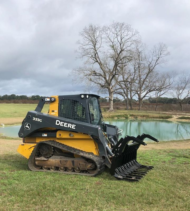 Yellow John Deere skid steer with grapple attachment in a grassy field near a pond.