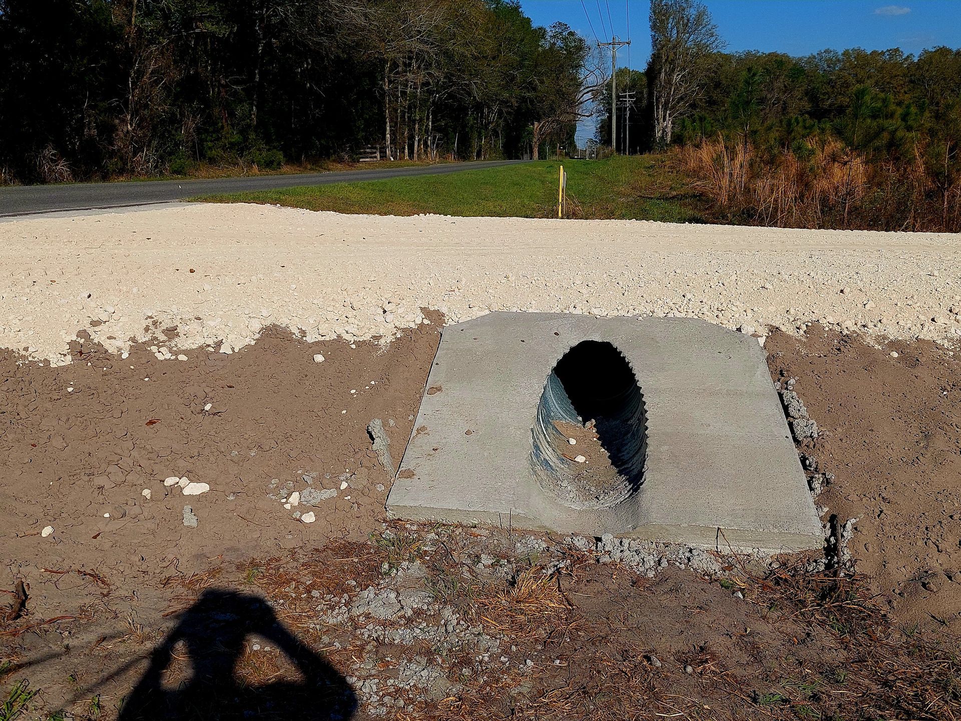 Concrete drainage culvert under a road with gravel shoulder, grass and trees in the background.