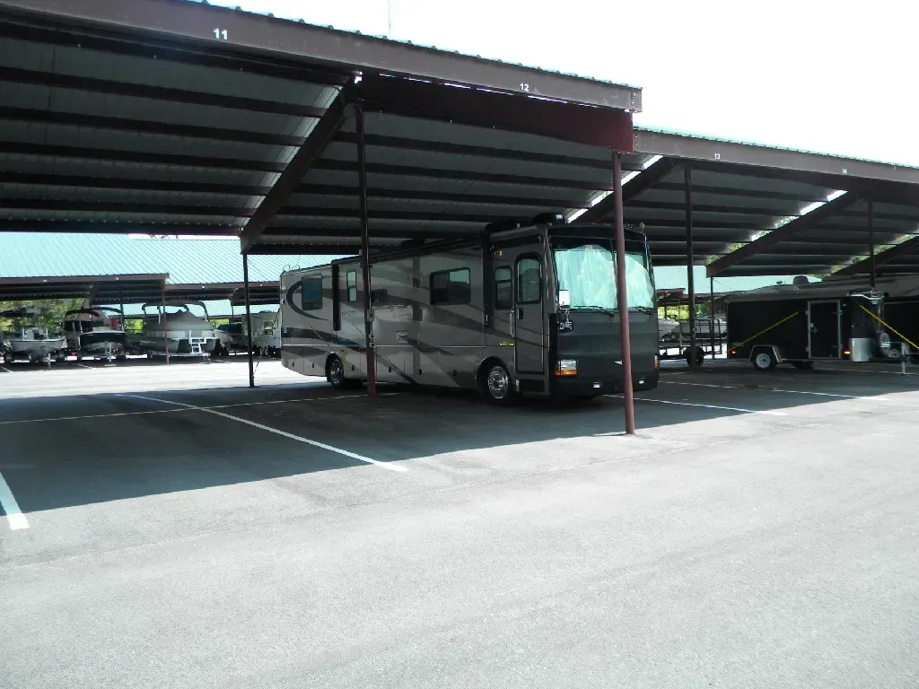 RV parked under a metal awning in a paved lot with other vehicles in the background.