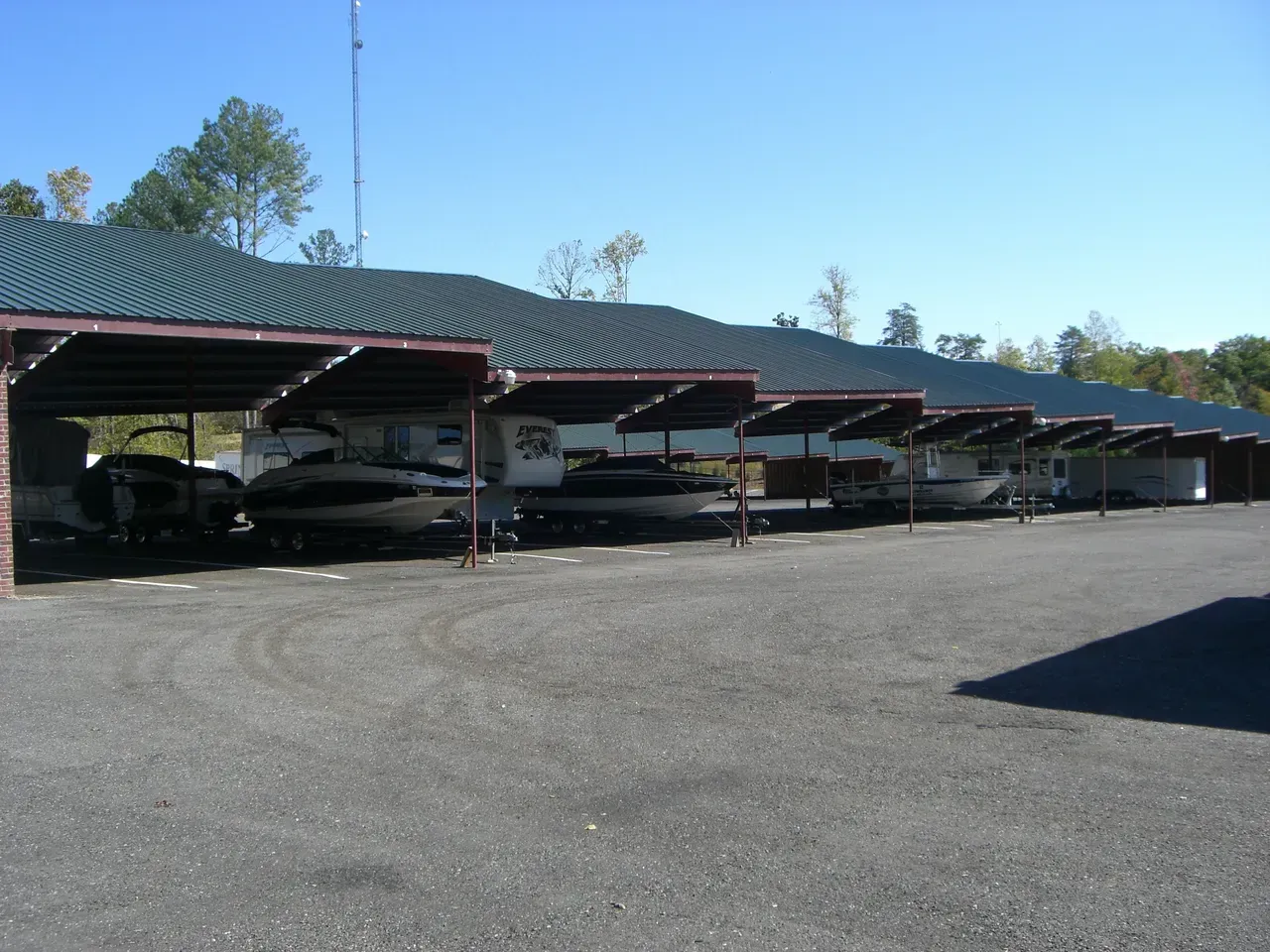 Cars parked under a long, covered structure with a green roof, on a gravel lot. Sunny day.