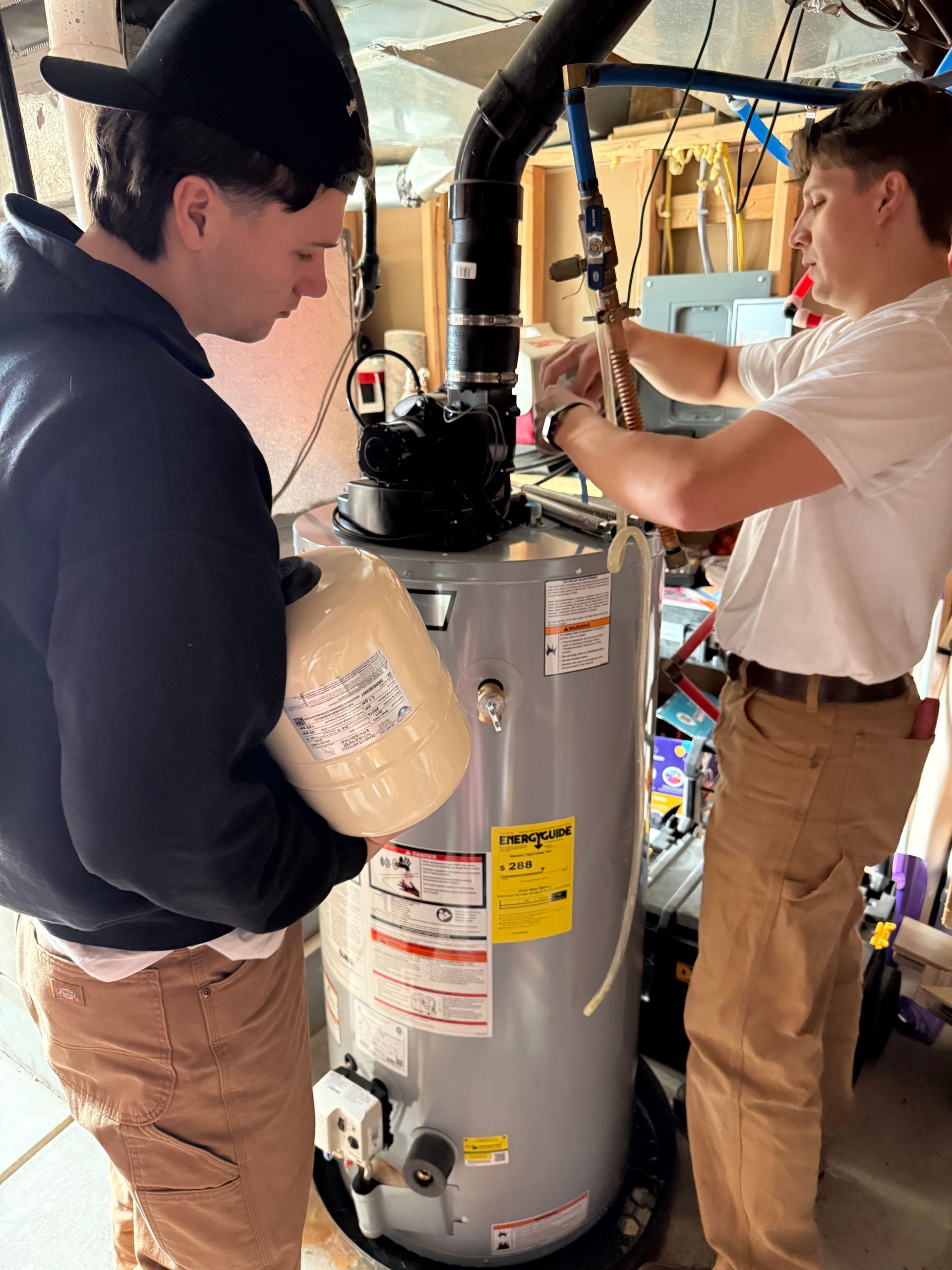 Two people work on a water heater in a garage, one holding a thermal expansion tank while the other adjusts plumbing pipes.