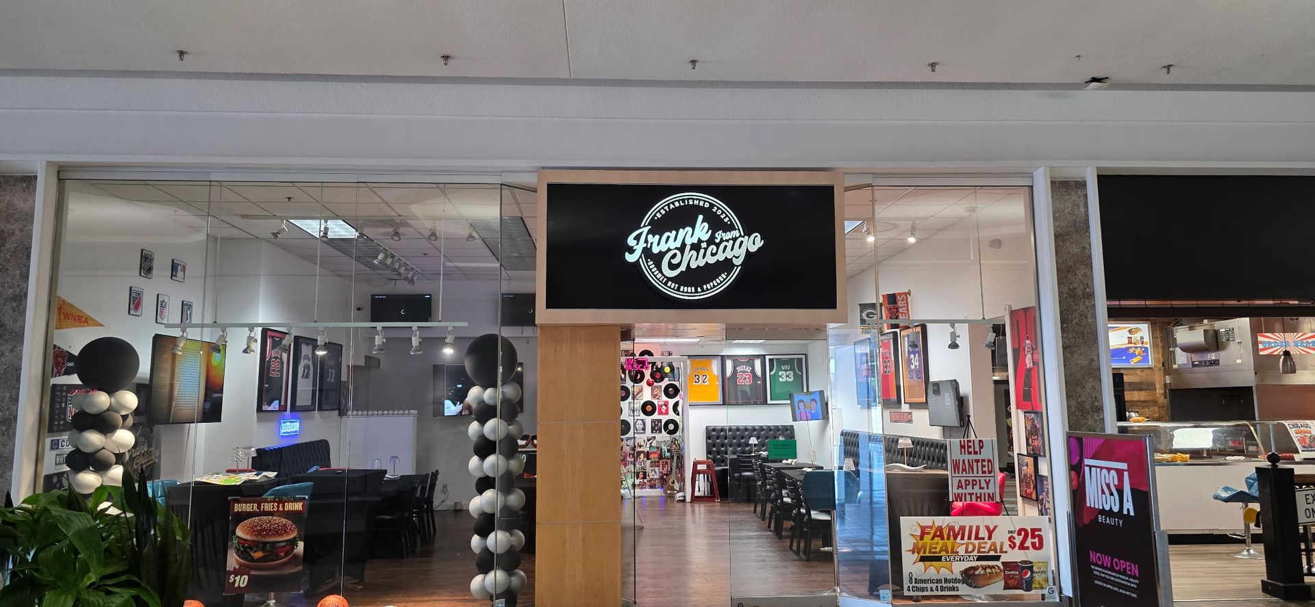 Storefront entrance with a black sign, bright interior lights, and shelves of products inside a mall corridor