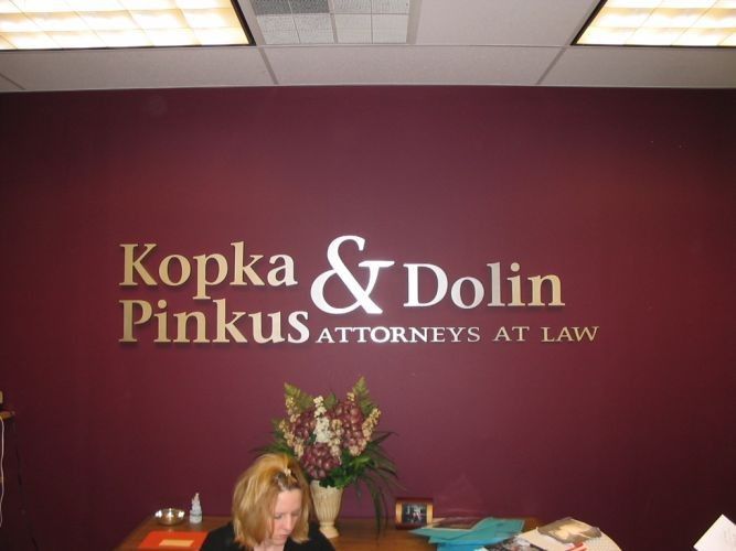 Gold lettering: Kopka & Dolin, Pinkus Attorneys At Law on a maroon wall, woman behind a desk.