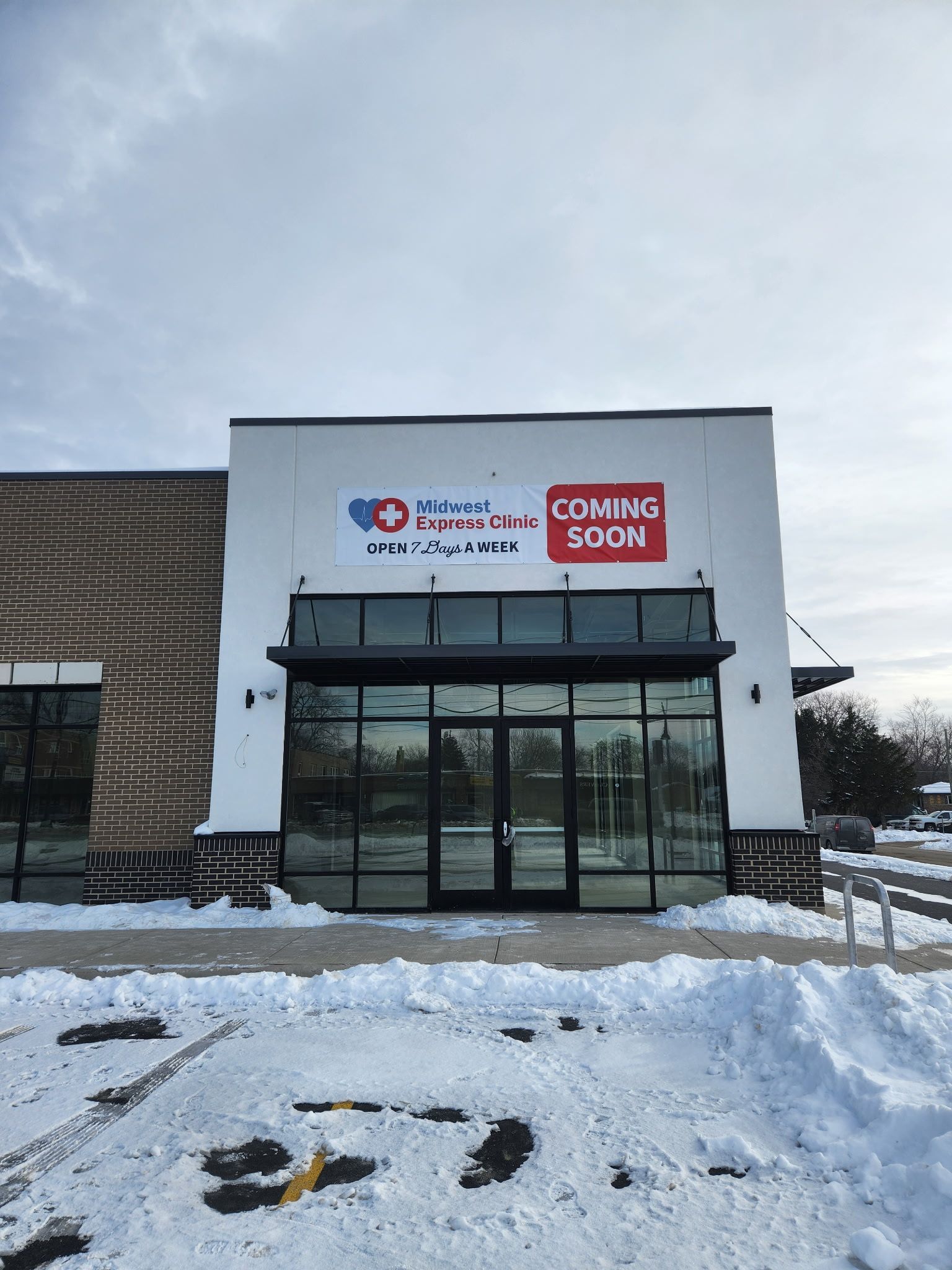 Snowy storefront with a glass entrance and red signs on a white building under a cloudy sky.