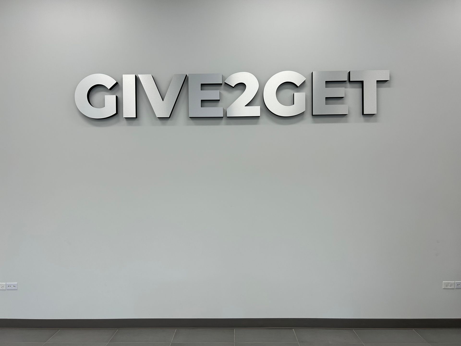White wall with metallic “GIVE2GET” letters and a dark floor trim.