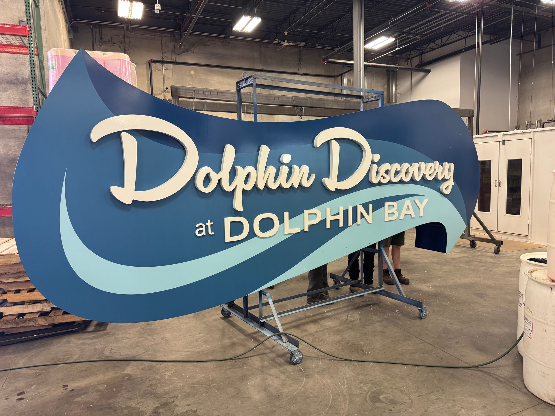 Blue wave-shaped “Dolphin Discovery at Dolphin Bay” sign in a workshop on a metal stand