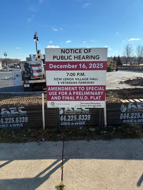 Public hearing notice sign on a roadside, with blue sky and parking lot in the background