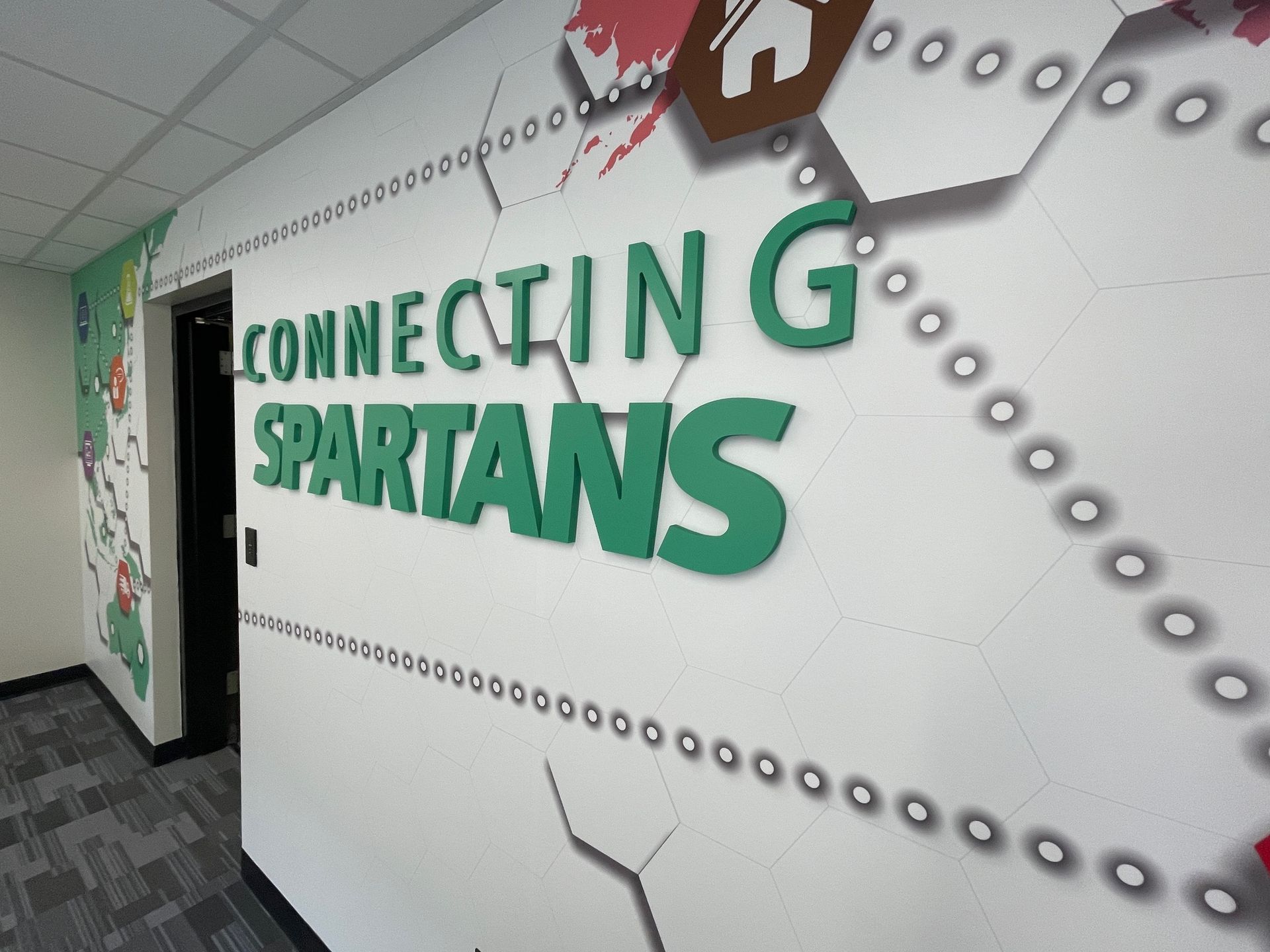 Hallway wall mural reading “Connecting Spartans” in green letters with dotted gray design and hanging icons