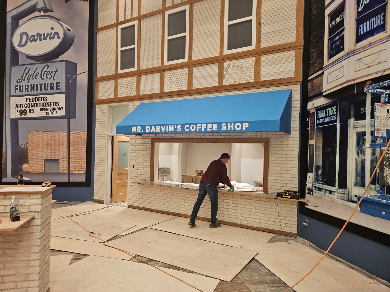 Man sweeping debris outside Darwin’s Coffee Shop on a damaged sidewalk, with boarded storefronts nearby