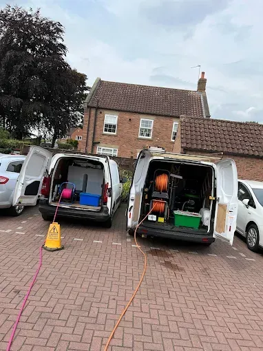 Two white vans parked, doors open, equipment visible. Cables extend to a bricked area. Building in background.