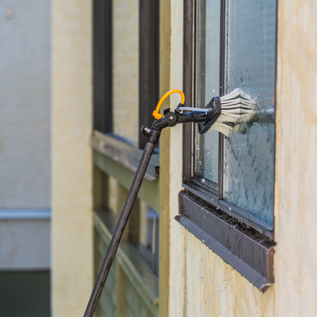 A person uses a long-handled window washing tool on a window next to a yellow exterior wall.