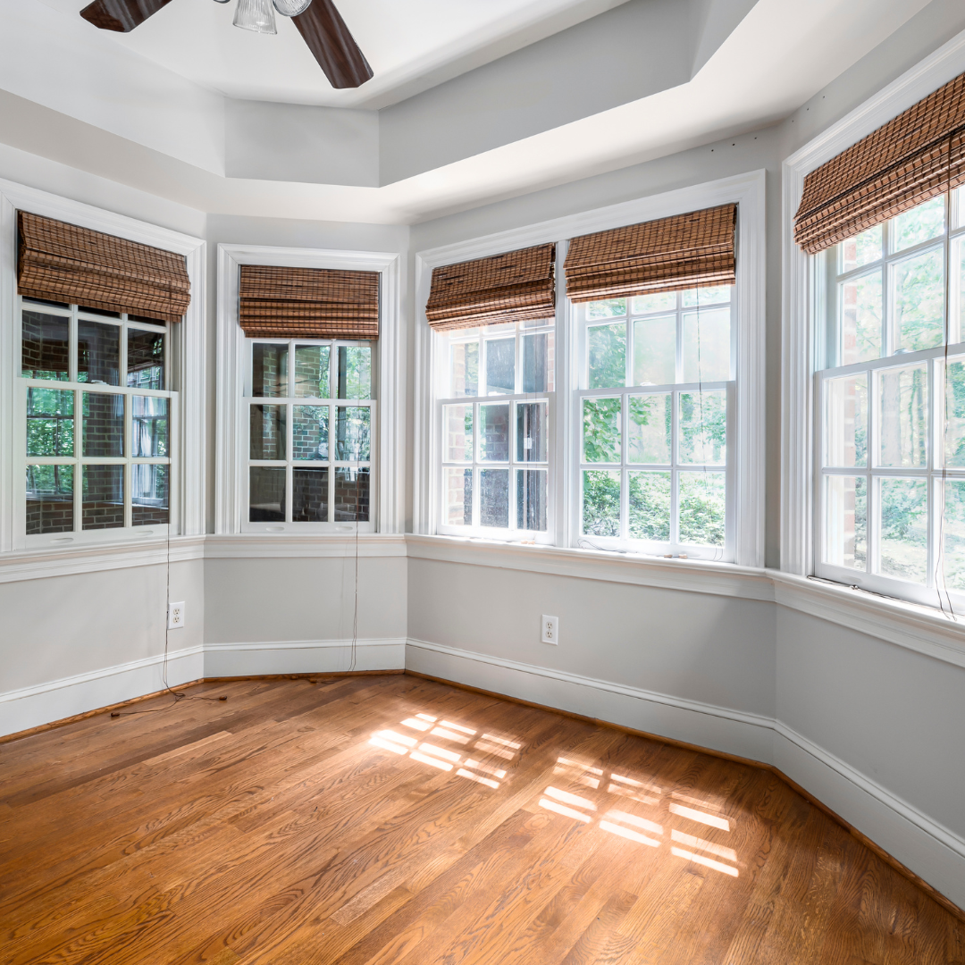 Empty room with hardwood floors, bay windows with woven shades, and a ceiling fan.