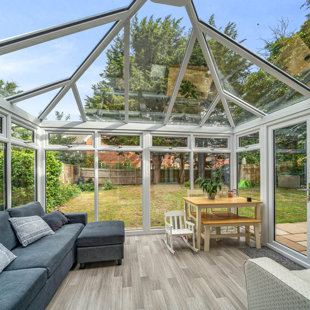Sunroom with glass walls and ceiling, gray sofa, small table, and view of a yard.