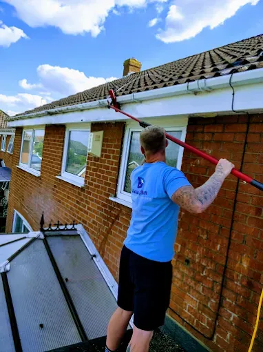 Man cleaning gutters on a brick house using a long-handled tool. Blue shirt, black shorts. Overcast sky.