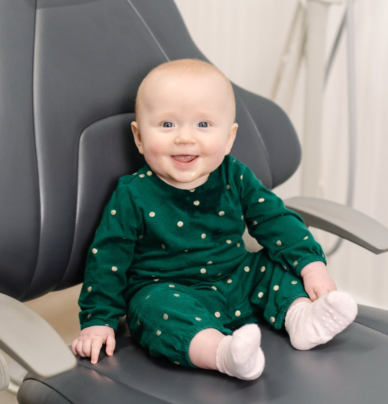 Smiling baby in a green polka-dot outfit sitting on a gray chair
